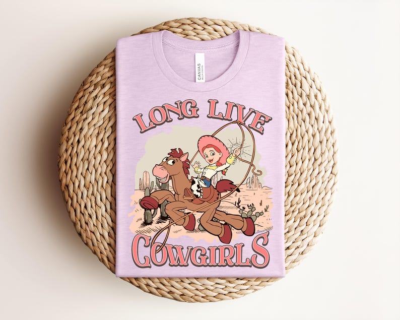 May include: A light purple t-shirt featuring a graphic of a cartoon cowgirl riding a brown horse in a desert landscape with cacti. The text "LONG LIVE COWGIRLS" is printed in a pink retro font above and below the illustration. The shirt has a crew neckline.