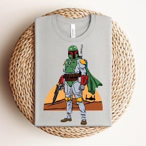 May include: A light gray t-shirt featuring a colorful illustration of a Star Wars character, Boba Fett. The character is depicted in his iconic green, red, and gray armor, with a green cape and a blaster. A sunset scene is in the background.