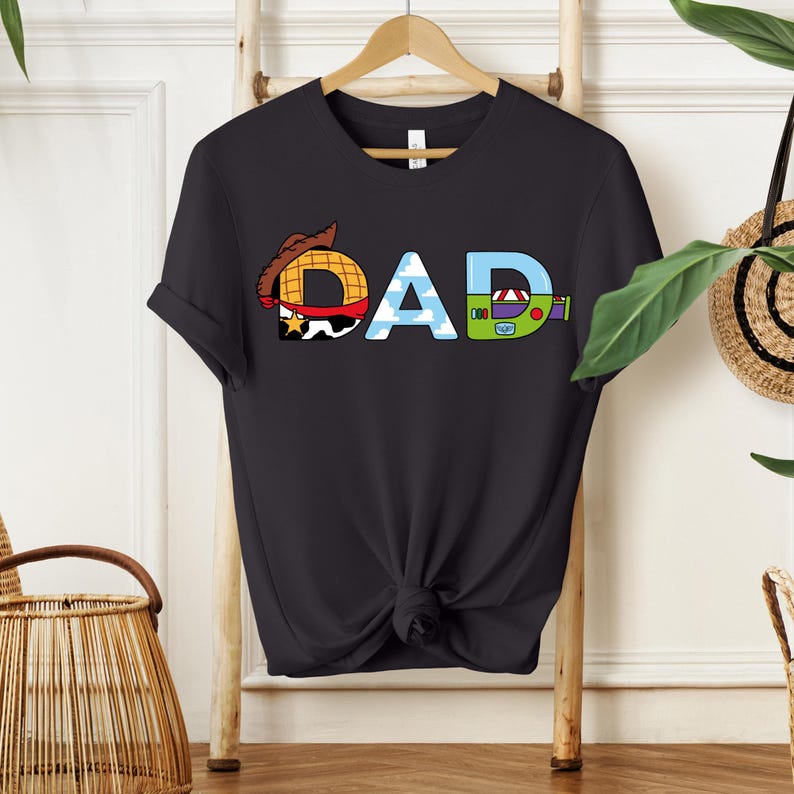 May include: Black t-shirt with the word "DAD" in a colorful, cartoon-style design. The letter "D" features a cowboy hat, the "A" has a blue sky with clouds, and the last "D" has a green and purple design.