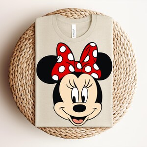 May include: A beige t-shirt featuring a graphic of Minnie Mouse's face. The design includes her black ears, a red bow with white polka dots, and a smiling face. The shirt is folded on a woven, circular surface.