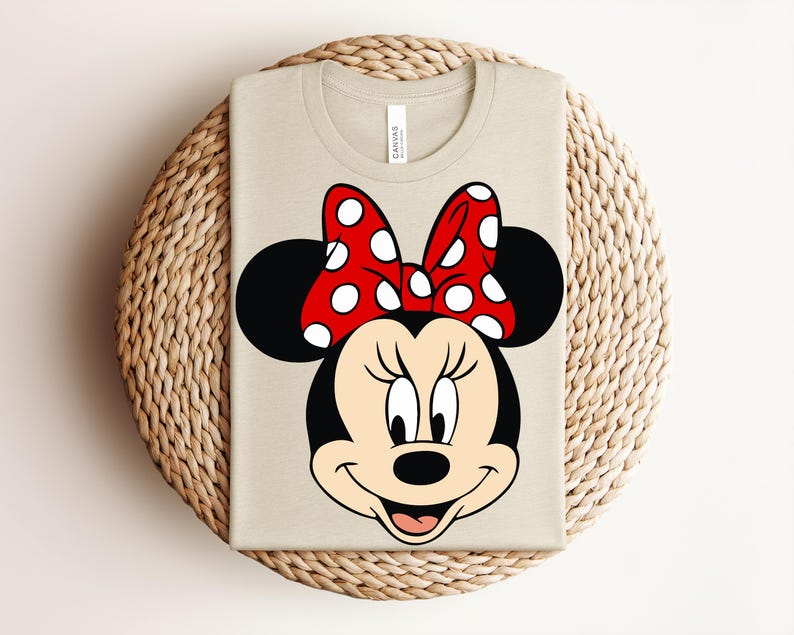 May include: A beige t-shirt featuring a graphic of Minnie Mouse's face. The design includes her black ears, a red bow with white polka dots, and a smiling face. The shirt is folded on a woven, circular surface.