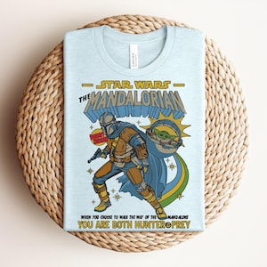 Puede incluir: Una camiseta azul claro con un gr&aacute;fico de The Mandalorian y Grogu. El dise&ntilde;o incluye el texto Star Wars The Mandalorian, Don't Hunt, y When You Choose To Walk The Way Of The Mandalore You Are Both Hunter And Prey en amarillo y azul.