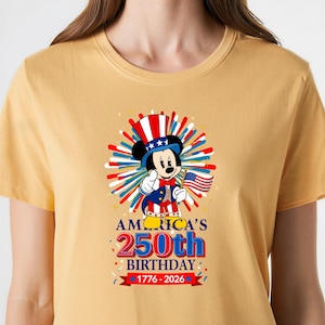 May include: Mustard yellow t-shirt featuring a patriotic Mickey Mouse design. Mickey is dressed in a red, white, and blue outfit, holding an American flag. The shirt celebrates America's 250th birthday, with text reading "America's 250th Birthday 1776-2026."