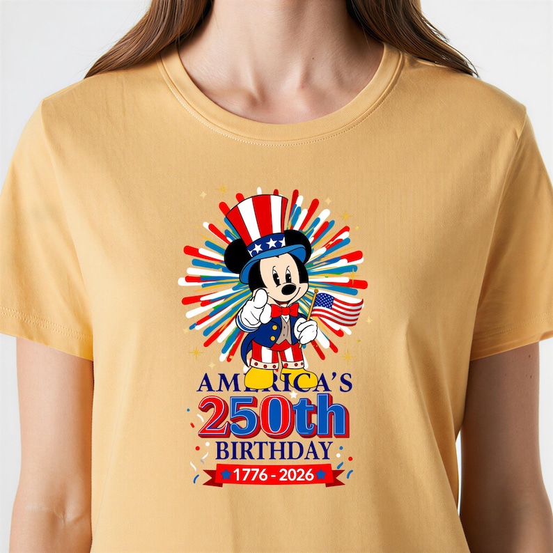 May include: Mustard yellow t-shirt featuring a patriotic Mickey Mouse design. Mickey is dressed in a red, white, and blue outfit, holding an American flag. The shirt celebrates America's 250th birthday, with text reading "America's 250th Birthday 1776-2026."
