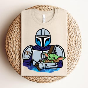Puede incluir: Camiseta beige con un gr&aacute;fico de un personaje mandaloriano sosteniendo a un alien&iacute;gena verde. El mandaloriano est&aacute; representado con una armadura plateada y azul, con el alien&iacute;gena acunado en sus brazos sobre un fondo estrellado. La camiseta est&aacute; doblada sobre una superficie tejida.