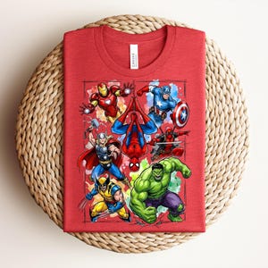 May include: A red t-shirt with a vibrant graphic of Marvel superheroes, such as Iron Man, Spider-Man, Captain America, Thor, Wolverine, Hulk, and Deadpool. The shirt is folded and placed on a woven, circular mat.