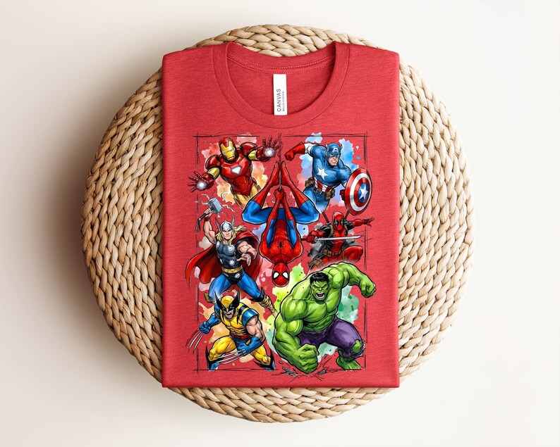 May include: A red t-shirt with a vibrant graphic of Marvel superheroes, such as Iron Man, Spider-Man, Captain America, Thor, Wolverine, Hulk, and Deadpool. The shirt is folded and placed on a woven, circular mat.