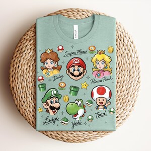 May include: A light green t-shirt featuring cartoon illustrations of Super Mario characters, including Mario, Luigi, Princess Peach, Daisy, Yoshi, and Toad. The words "Super Mario", "Daisy", "Princess Peach", "Luigi", "Yoshi", and "Toad" are written on the shirt.