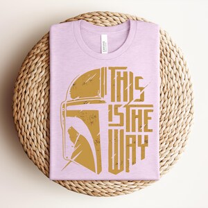 May include: A light purple crew neck t-shirt featuring a distressed gold graphic of a helmet silhouette on the left and the text THIS IS THE WAY stacked vertically on the right. The shirt has a white tag at the inner neckline with brand text.