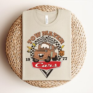 May include: A beige t-shirt featuring a graphic of the character Tow Mater from the movie Cars. The design includes the text Tow Mater, the number 1977, a Route 66 sign, a trophy, checkered flags, and the Cars logo in red and white.