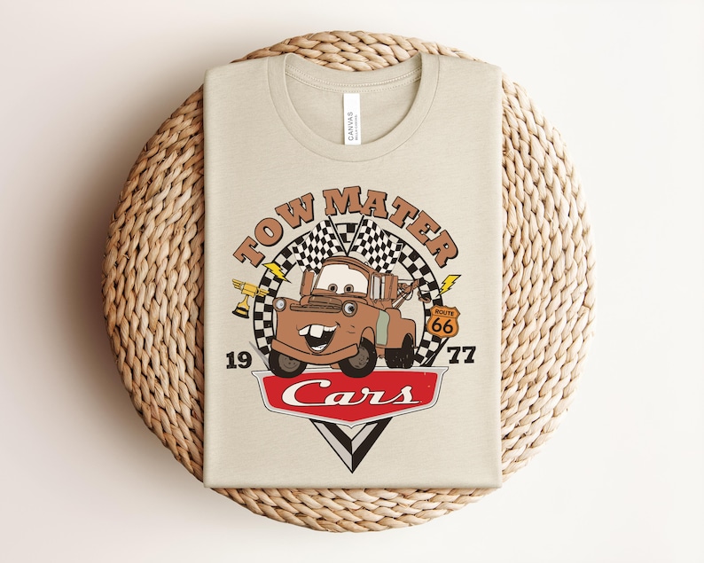 May include: A beige t-shirt featuring a graphic of the character Tow Mater from the movie Cars. The design includes the text Tow Mater, the number 1977, a Route 66 sign, a trophy, checkered flags, and the Cars logo in red and white.