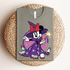 May include: A light olive green t-shirt featuring a cartoon Minnie Mouse dressed as a witch. She wears a purple dress and hat with a red bow, holding a wand. The design includes potion bottles, a crystal ball, and open books.