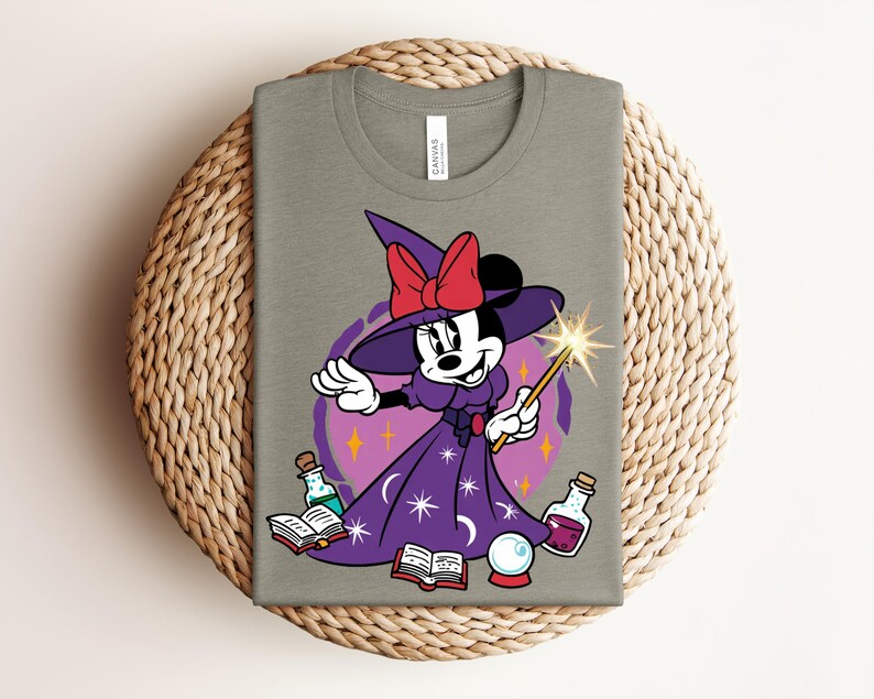 May include: A light olive green t-shirt featuring a cartoon Minnie Mouse dressed as a witch. She wears a purple dress and hat with a red bow, holding a wand. The design includes potion bottles, a crystal ball, and open books.
