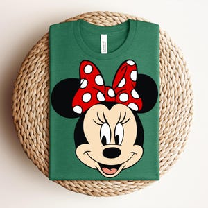 May include: A green t-shirt featuring a graphic of Minnie Mouse's face. The design includes her black ears, a red bow with white polka dots, and a smiling face. The shirt is folded and placed on a woven mat.