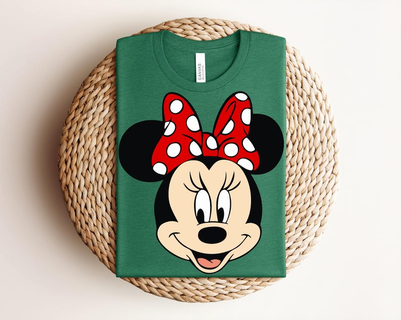 May include: A green t-shirt featuring a graphic of Minnie Mouse's face. The design includes her black ears, a red bow with white polka dots, and a smiling face. The shirt is folded and placed on a woven mat.