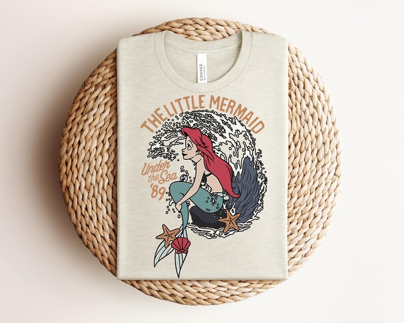 May include: A cream-colored t-shirt featuring a graphic of a mermaid with red hair and a teal tail sitting on a rock. The text reads THE LITTLE MERMAID, Under the Sea, and '89. The design includes a large wave, two starfish, and a pink seashell.