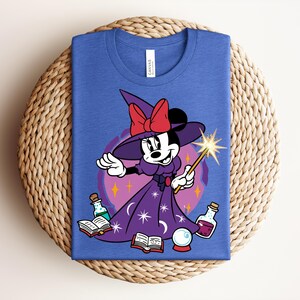 May include: Blue t-shirt featuring a cartoon Minnie Mouse dressed as a witch, holding a wand with a starburst. She wears a purple dress and hat with a red bow. The design includes potion bottles, a crystal ball, and open books.