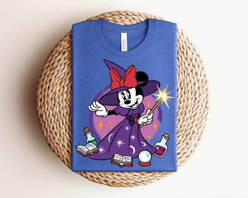 May include: Blue t-shirt featuring a cartoon Minnie Mouse dressed as a witch, holding a wand with a starburst. She wears a purple dress and hat with a red bow. The design includes potion bottles, a crystal ball, and open books.