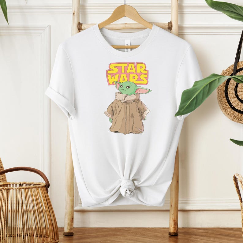 Puede incluir: Camiseta blanca con un gr&aacute;fico de un personaje alien&iacute;gena verde de Star Wars, con un abrigo marr&oacute;n. El texto "STAR WARS" est&aacute; encima del personaje en amarillo y rosa. La camiseta est&aacute; colgada en una percha de madera.
