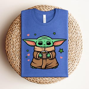 May include: A blue t-shirt featuring a cartoon character with large green ears, holding a cup. The character is wearing a brown coat and is surrounded by hearts and stars. The shirt is folded on a woven surface.