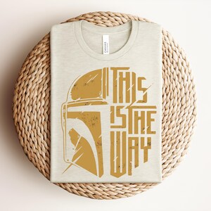 May include: A cream-colored t-shirt featuring a distressed gold graphic of a helmet and the text "THIS IS THE WAY" printed on the front. The design is centered on the chest, showcasing a stylized warrior mask alongside bold, blocky lettering.
