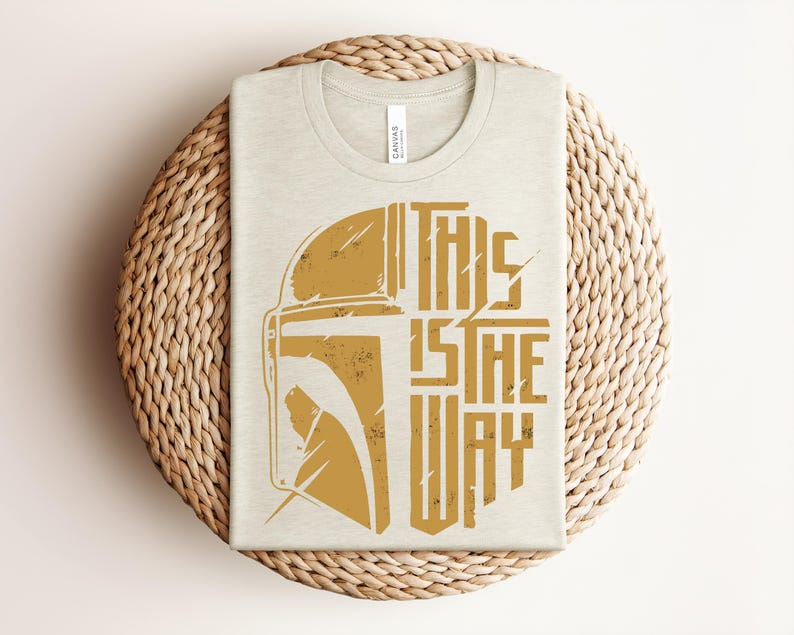 May include: A cream-colored t-shirt featuring a distressed gold graphic of a helmet and the text "THIS IS THE WAY" printed on the front. The design is centered on the chest, showcasing a stylized warrior mask alongside bold, blocky lettering.