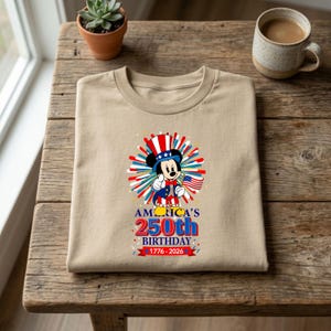 May include: A tan t-shirt featuring a graphic of Mickey Mouse in patriotic attire, holding a small American flag. The design includes the text "America's 250th Birthday 1776-2026" in red, white, and blue.