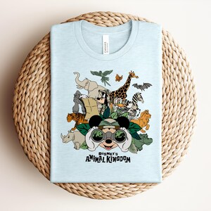Puede incluir: Una camiseta azul claro con un estampado gr&aacute;fico de personajes de Disney, incluyendo a Mickey Mouse mirando a trav&eacute;s de binoculares, rodeado de animales como un elefante, jirafa, tigre y cebra, con el texto Disney's ANIMAL KINGDOM.
