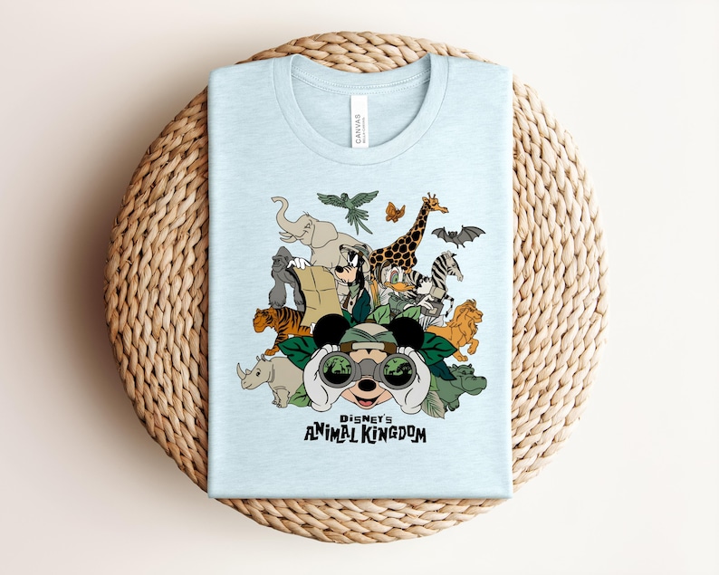 Puede incluir: Una camiseta azul claro con un estampado gr&aacute;fico de personajes de Disney, incluyendo a Mickey Mouse mirando a trav&eacute;s de binoculares, rodeado de animales como un elefante, jirafa, tigre y cebra, con el texto Disney's ANIMAL KINGDOM.