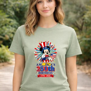 May include: Sage green t-shirt featuring a graphic of Mickey Mouse in patriotic attire, holding a small American flag. The design includes text that reads "America's 250th Birthday 1776-2026" in red, white, and blue.