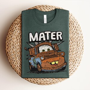 May include: A dark green crew neck t-shirt featuring a graphic print of a rusty brown tow truck character with large eyes and buck teeth. The word MATER is printed in large white block letters above the truck, which is set against a black starburst.