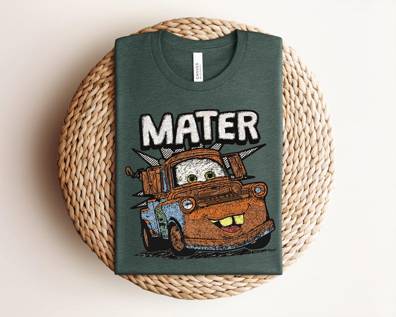 May include: A dark green crew neck t-shirt featuring a graphic print of a rusty brown tow truck character with large eyes and buck teeth. The word MATER is printed in large white block letters above the truck, which is set against a black starburst.