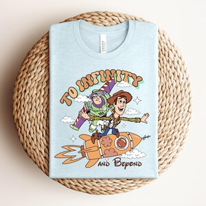 May include: A light blue t-shirt featuring a graphic of Buzz Lightyear and Woody from Toy Story. Woody rides an orange rocket while Buzz flies behind him. The text reads TO INFINITY AND BEYOND in a retro font, surrounded by clouds and stars.