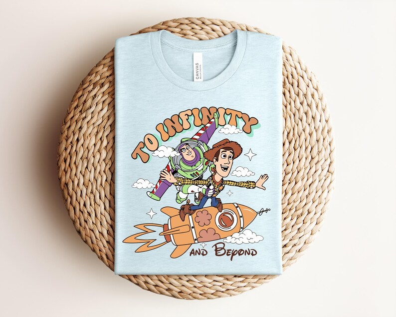 May include: A light blue t-shirt featuring a graphic of Buzz Lightyear and Woody from Toy Story. Woody rides an orange rocket while Buzz flies behind him. The text reads TO INFINITY AND BEYOND in a retro font, surrounded by clouds and stars.