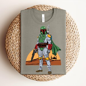 May include: A folded olive green t-shirt featuring a colorful illustration of a Star Wars character, Boba Fett. The character is depicted in his iconic green, red, and gray armor, holding a blaster. A desert landscape and a setting sun are in the background.