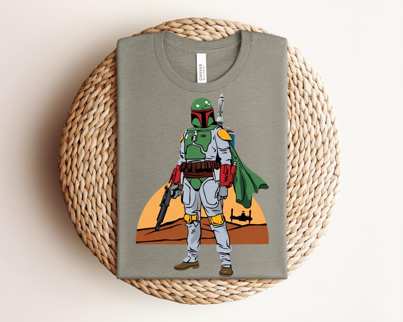 May include: A folded olive green t-shirt featuring a colorful illustration of a Star Wars character, Boba Fett. The character is depicted in his iconic green, red, and gray armor, holding a blaster. A desert landscape and a setting sun are in the background.