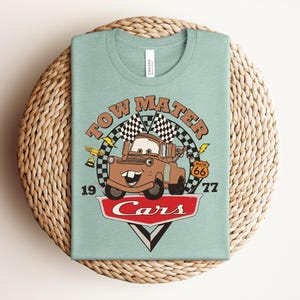 May include: A light green t-shirt featuring a graphic of the character Tow Mater from the movie Cars. The design includes the text Tow Mater, 1977, Route 66, and Cars, along with checkered flags, a gold trophy, and lightning bolt accents.