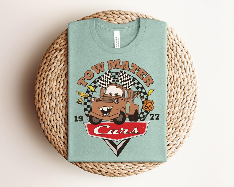 May include: A light green t-shirt featuring a graphic of the character Tow Mater from the movie Cars. The design includes the text Tow Mater, 1977, Route 66, and Cars, along with checkered flags, a gold trophy, and lightning bolt accents.