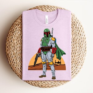 May include: A light purple t-shirt featuring a graphic of a Star Wars character in green, gray, and red armor, with a green cape and a blaster. The design includes a desert landscape with a setting sun and a spaceship silhouette.