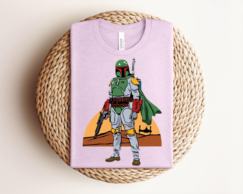 May include: A light purple t-shirt featuring a graphic of a Star Wars character in green, gray, and red armor, with a green cape and a blaster. The design includes a desert landscape with a setting sun and a spaceship silhouette.