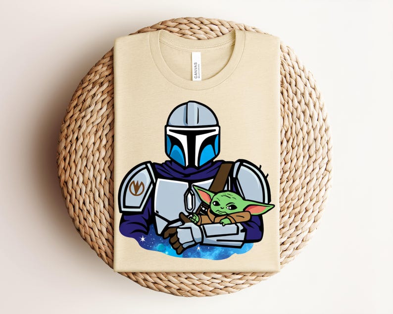 Puede incluir: Camiseta beige con una ilustraci&oacute;n de dibujos animados de un personaje con armadura plateada y azul sosteniendo una peque&ntilde;a criatura verde. El dise&ntilde;o incluye un fondo azul y blanco con tem&aacute;tica espacial. La camiseta est&aacute; doblada sobre una superficie tejida.