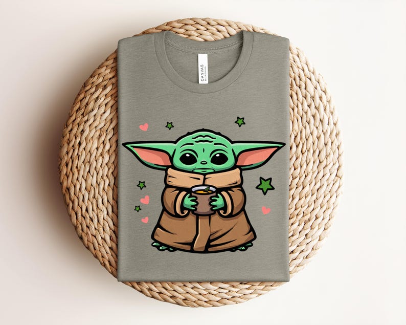 May include: A sage green t-shirt featuring a cartoon character holding a cup. The character has large green ears, big eyes, and is wearing a brown coat. Pink hearts and green stars surround the character. The shirt is folded on a woven mat.