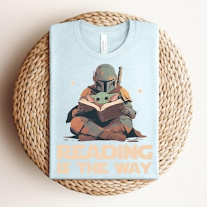 May include: A light blue t-shirt featuring a graphic of a helmeted character in armor sitting cross-legged and reading a book with a small green creature. The text "READING IS THE WAY" is printed in large, light orange letters below the illustration.