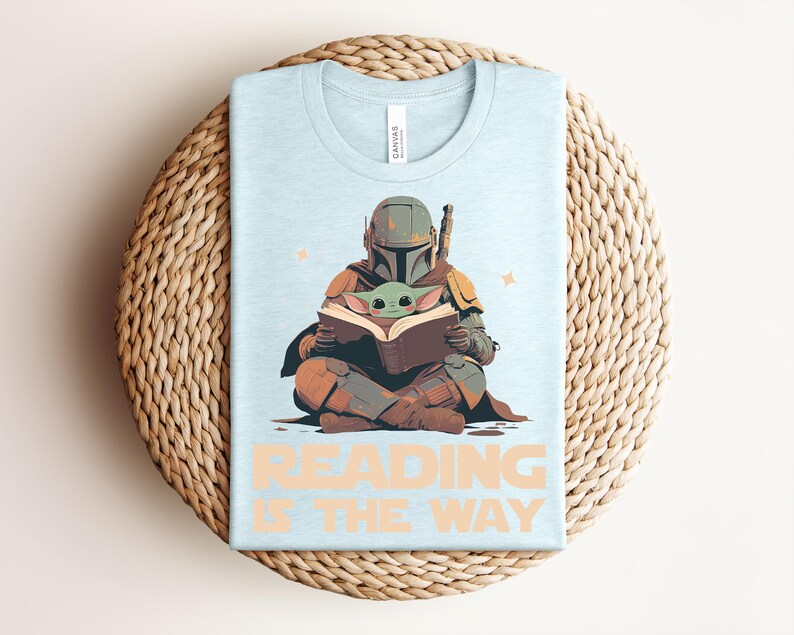 May include: A light blue t-shirt featuring a graphic of a helmeted character in armor sitting cross-legged and reading a book with a small green creature. The text "READING IS THE WAY" is printed in large, light orange letters below the illustration.