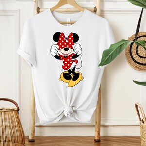 May include: White t-shirt featuring a cartoon illustration of Minnie Mouse. Minnie is depicted in a red polka-dot dress and bow, with yellow shoes. The shirt is hanging on a wooden rack.