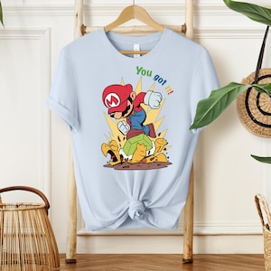 May include: Light blue t-shirt featuring a cartoon illustration of a character in a red hat and overalls, punching forward. The text "You got it!" is above the character. The character is standing on a turtle shell.