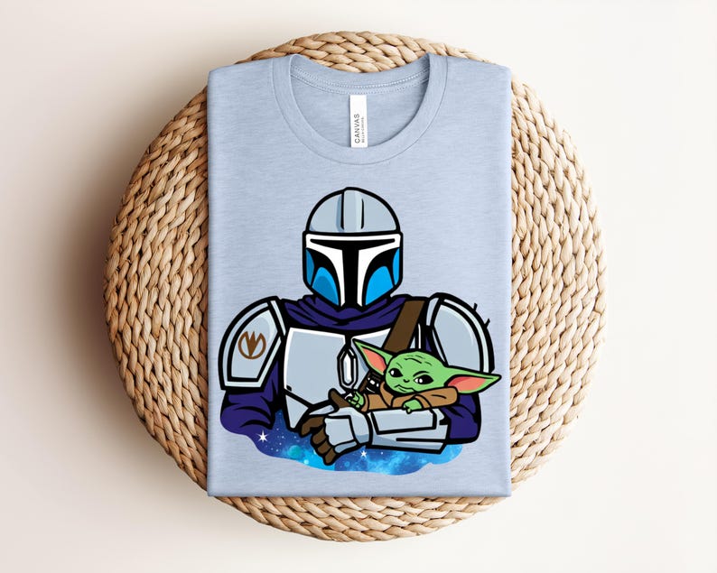Puede incluir: Camiseta azul claro con una ilustraci&oacute;n de dibujos animados de un personaje mandaloriano sosteniendo a un alien&iacute;gena verde. El dise&ntilde;o incluye un casco, armadura y un fondo tem&aacute;tico espacial. La camiseta est&aacute; doblada sobre una superficie tejida.