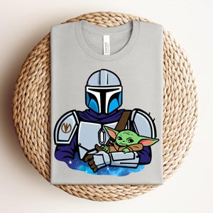 Puede incluir: Una camiseta gris claro con una ilustraci&oacute;n de dibujos animados de un personaje mandaloriano sosteniendo a un alien&iacute;gena verde. El mandaloriano lleva un casco y una armadura con detalles azules y plateados. El alien&iacute;gena es peque&ntilde;o y verde, con orejas grandes.