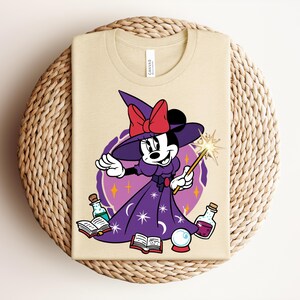 May include: Beige t-shirt featuring a cartoon Minnie Mouse dressed as a witch, wearing a purple dress and hat with a red bow. She holds a wand with a starburst, surrounded by potion bottles, books, and a crystal ball. The shirt has a "CANVAS" tag.