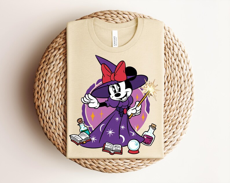 May include: Beige t-shirt featuring a cartoon Minnie Mouse dressed as a witch, wearing a purple dress and hat with a red bow. She holds a wand with a starburst, surrounded by potion bottles, books, and a crystal ball. The shirt has a "CANVAS" tag.